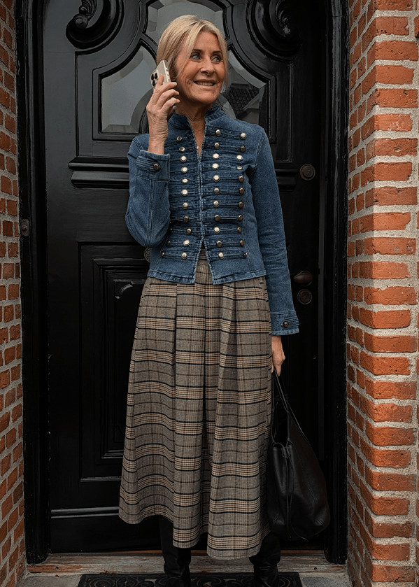 En kvinde står foran en sort dør og taler i mobiltelefonen. Hun er iført en militærinspireret jakke i denim, en midi-nederdel i ternet stof og en skuldertaske. - Fredags outfittet
