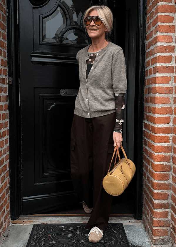 En kvinde iført en beige cardigan over en mønstret bluse, mørke bukser og store solbriller står i en døråbning og holder en beige, cylindrisk håndtaske.