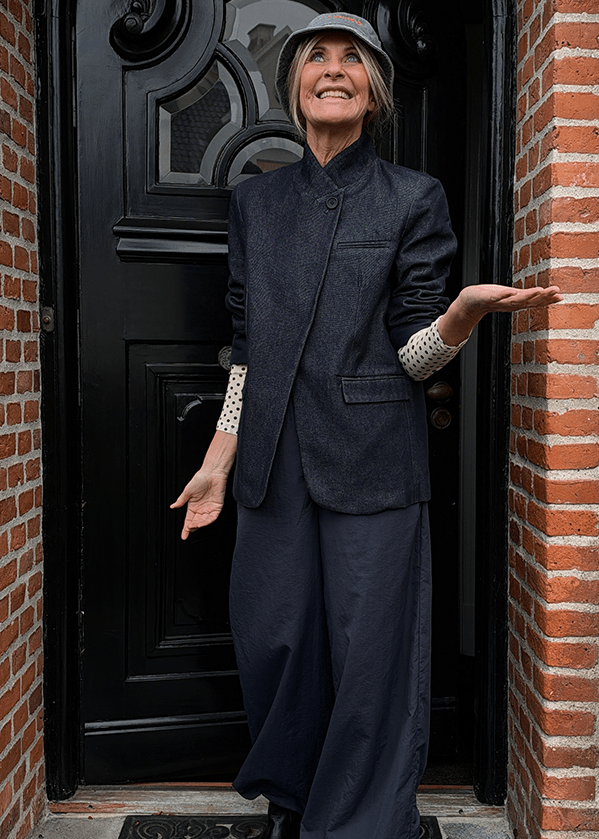 En kvinde iført en bucket hat, en mørk blazer og bukser med brede ben står foran en sort dør og gestikulerer med hænderne. - Tirsdags outfittet