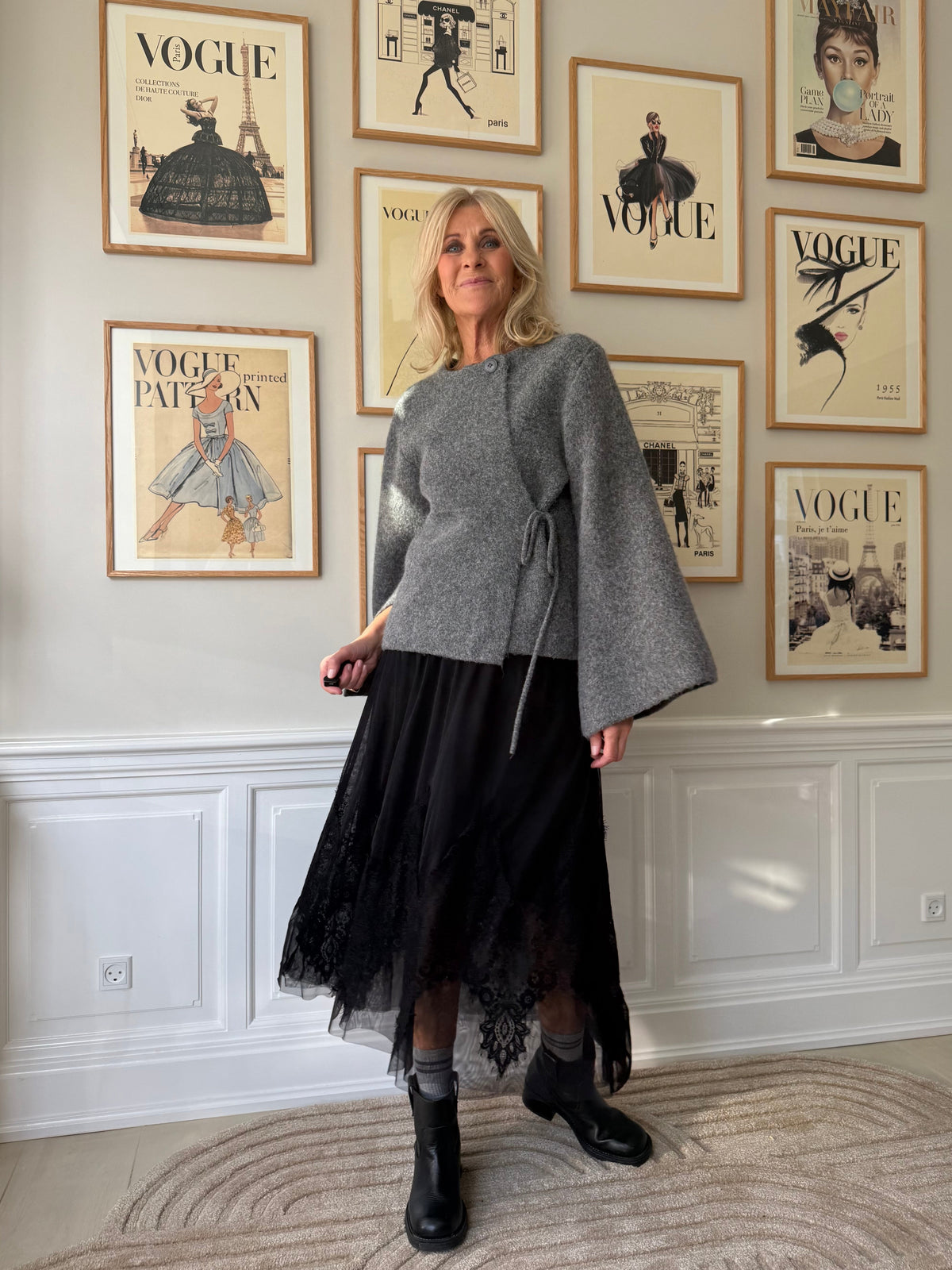 En kvinde står inde i et rum iført en grå wrap-sweater, en sort blonderkjole og sorte støvler. Bag hende hænger indrammede forsider fra magasinet Vogue i et gittermønster på en cremefarvet væg.