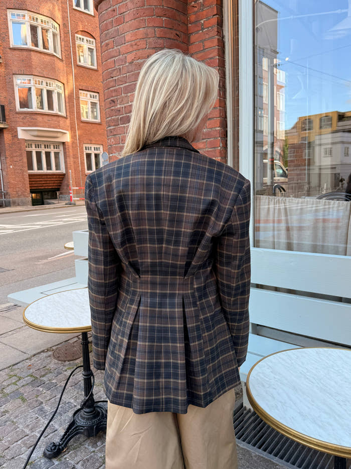 En person med langt blondt hår, iført en brun ternet blazer og beige bukser, står udenfor ved nogle runde caféborde på et brostensbelagt fortov og har ryggen til kameraet. - Ternet blazer