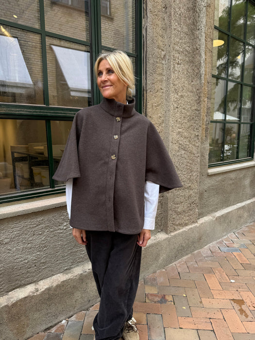 En kvinde står udenfor ved et stort vindue iført en brun poncho med knapper over en hvid skjorte og mørke bukser. Hun kigger til siden og smiler let. - Kappe