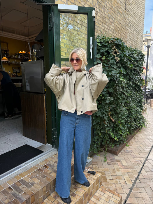 En person iført solbriller, en beige jakke med brede ærmer og blå jeans med brede ben står uden for en café med murstensvægge og grønne planter. - Kort trenchcoat