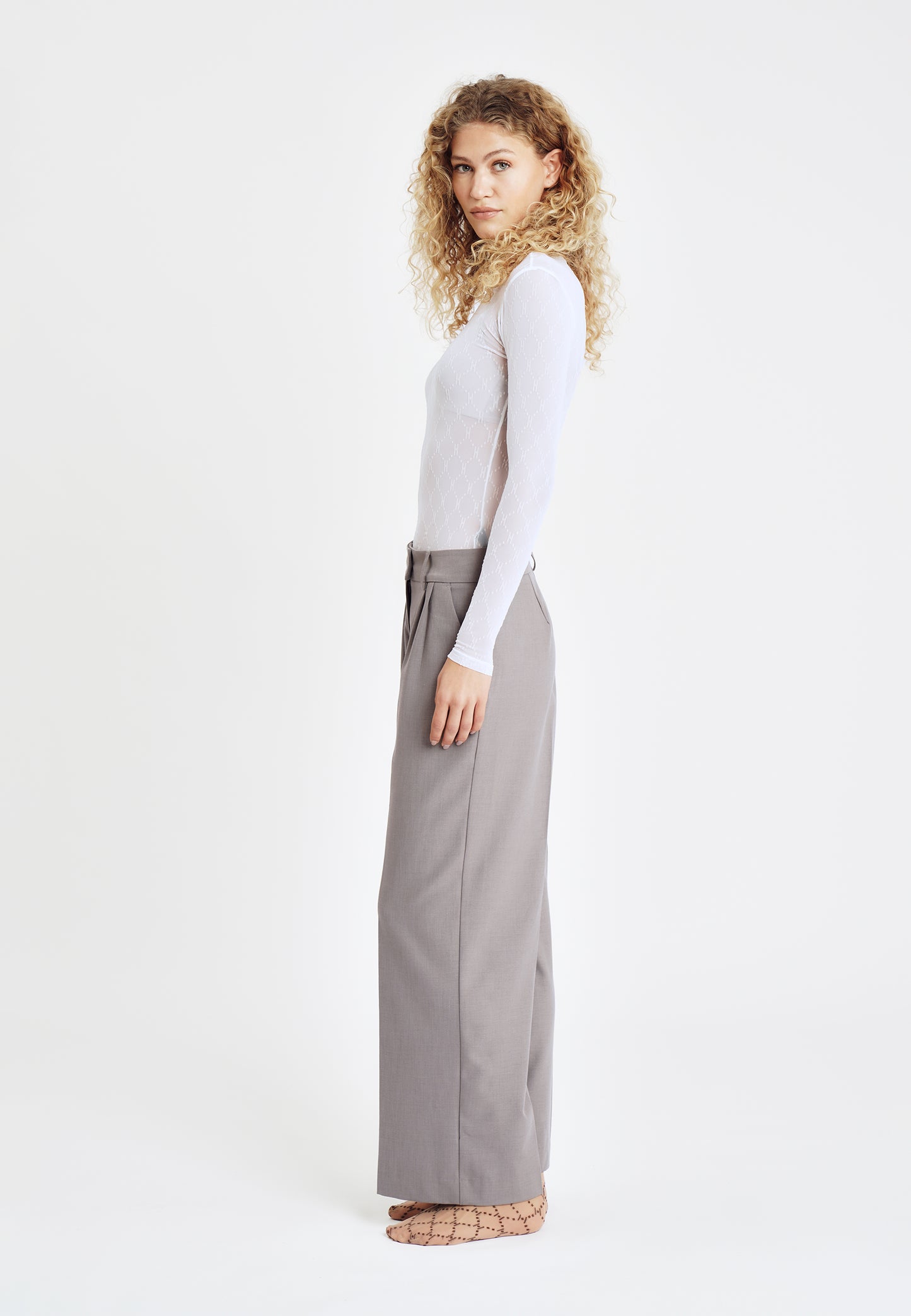 Kvinde, der står med siden til, iført en HYPE THE DETAIL-meshbluse i offwhite, grå bukser med brede ben og brune mønstrede sko.