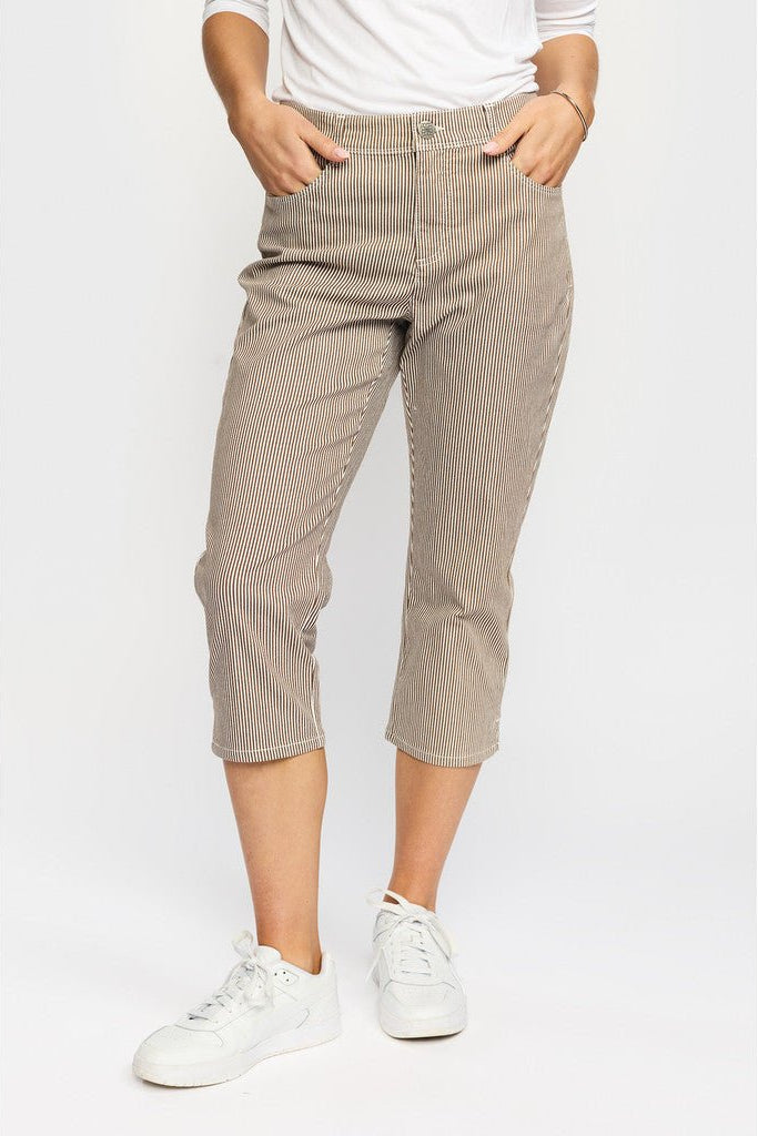 En person har 2-Biz Fira Brun-bukser, hvide sneakers og en hvid skjorte på, mens vedkommende står foran en ensfarvet baggrund.