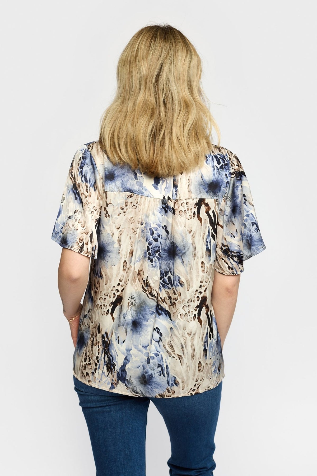 En person iført en 2-Biz Amora-bluse med blåt mønster, med lyst, bølget hår og blå jeans.
