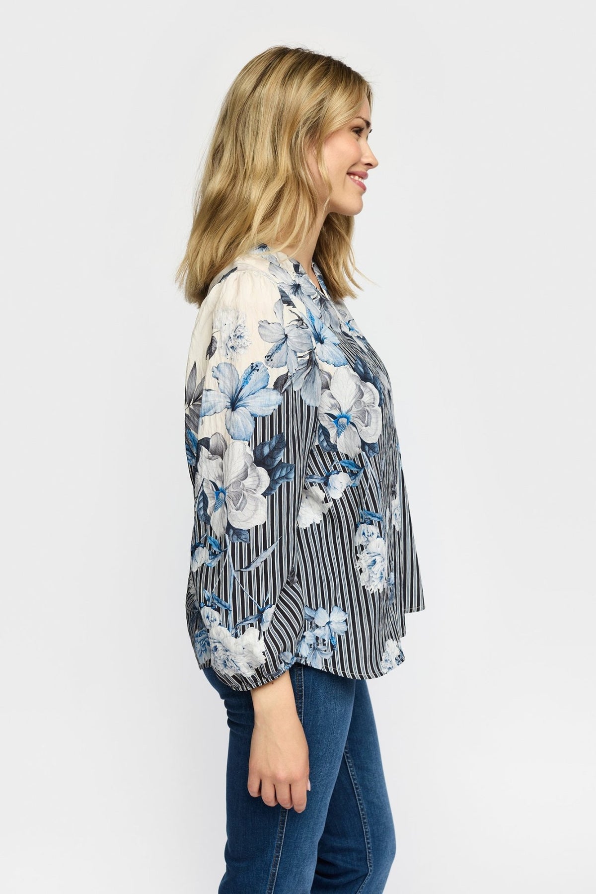 En kvinde set i profil er iført en 2-Biz Jana-bluse med blåt mønster og jeans og står foran en ensfarvet baggrund.