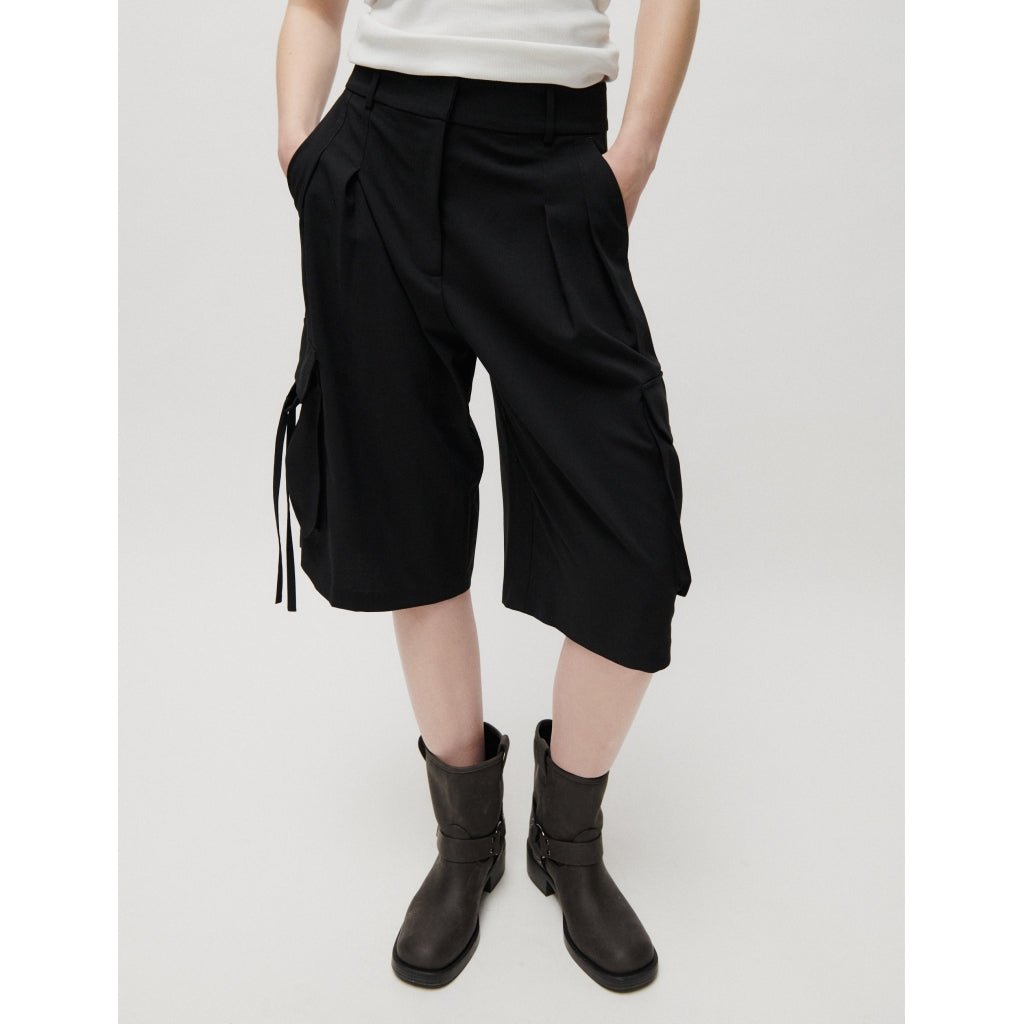 En person iført 2NDDAY Hattori Sort-shorts, en hvid top og sorte støvler står foran en ensfarvet lys baggrund.