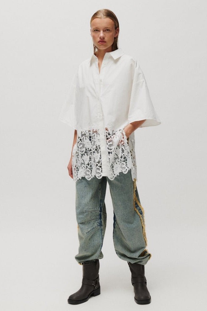 En person iført en Edith Off White-skjorte fra 2NDDAY, løse jeans og sorte støvler står foran en ensfarvet baggrund.
