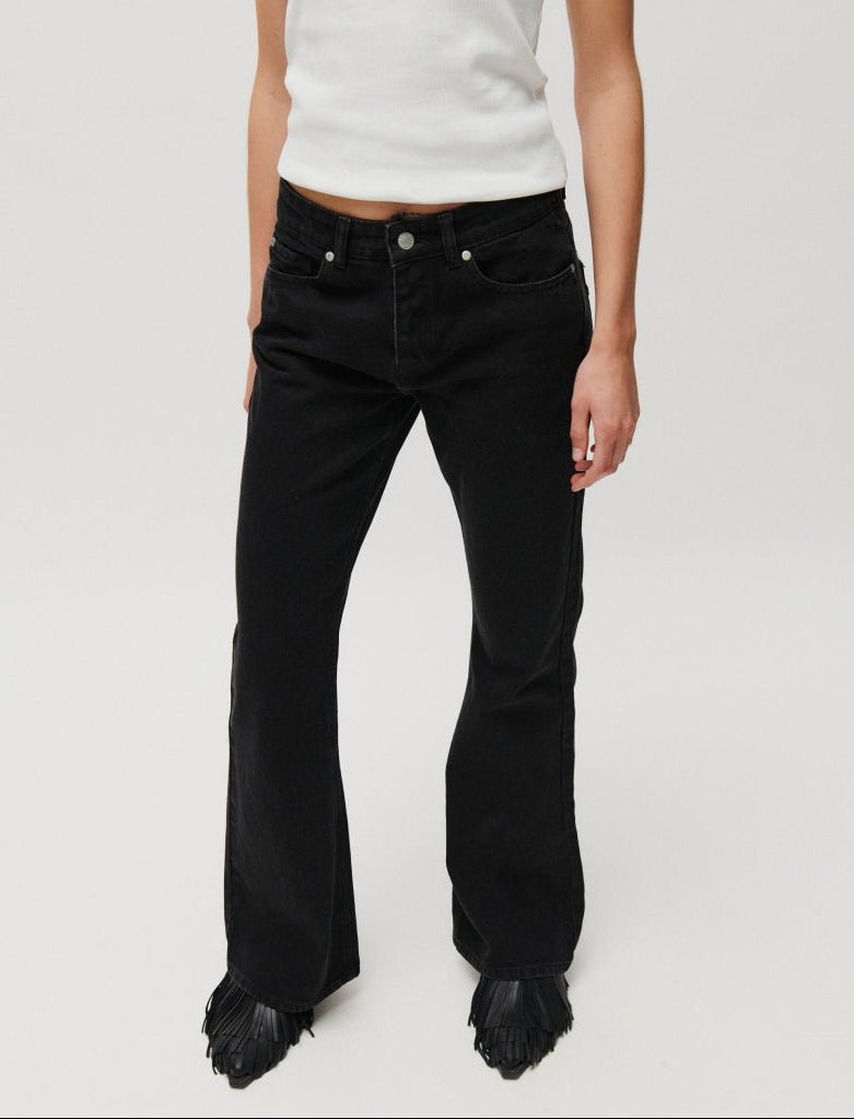 En person iført et par 2NDDAY Lamar Sort-jeans, en hvid ærmeløs top og sorte sko står foran en ensfarvet baggrund.