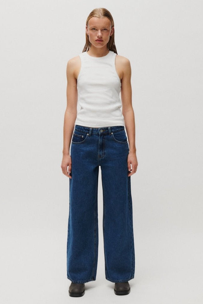 En person står med ansigtet vendt fremad iført en hvid, ærmeløs top og 2NDDAY Roden Denim-jeans samt sorte sko foran en ensfarvet baggrund.