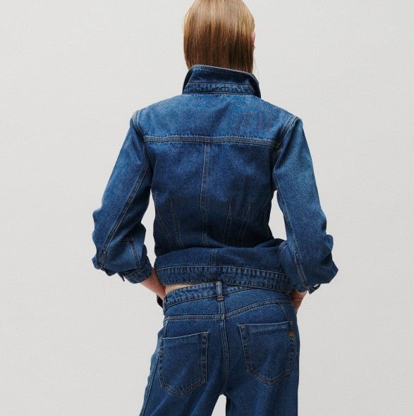 En person med glat hår har en 2NDDAY Billa Denim-truckerjakke og jeans på, står med hænderne i siden og vender ryggen til.