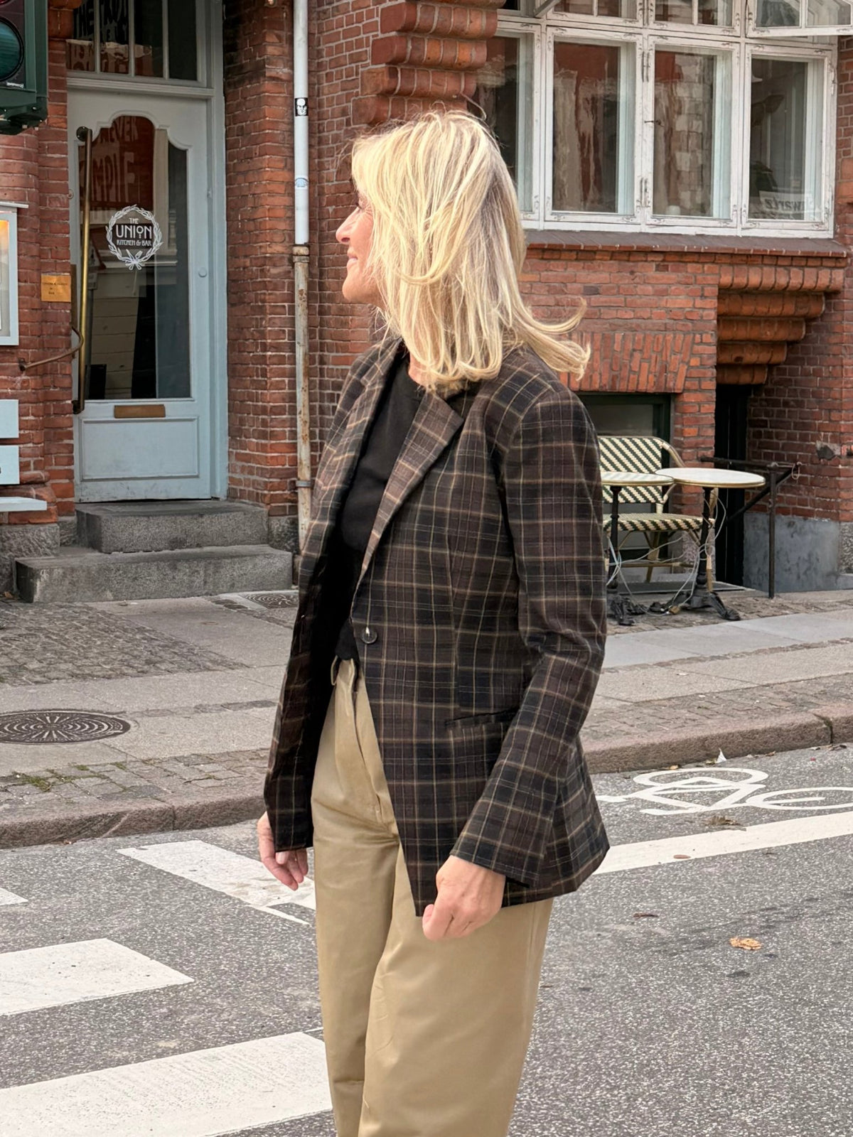En kvinde med blondt hår, iført en ternet blazer fra 2NDDAY 2NDMaeva Ternet og kakifarvede bukser, står ved en café på en gade i byen.