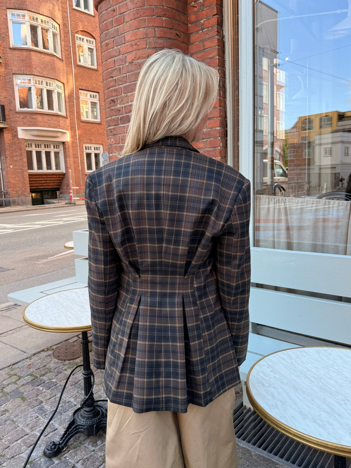 En person med langt blondt hår iført en 2NDDAY 2NDMaeva Ternet-blazer i tern står udenfor ved runde borde og murstensbygninger.