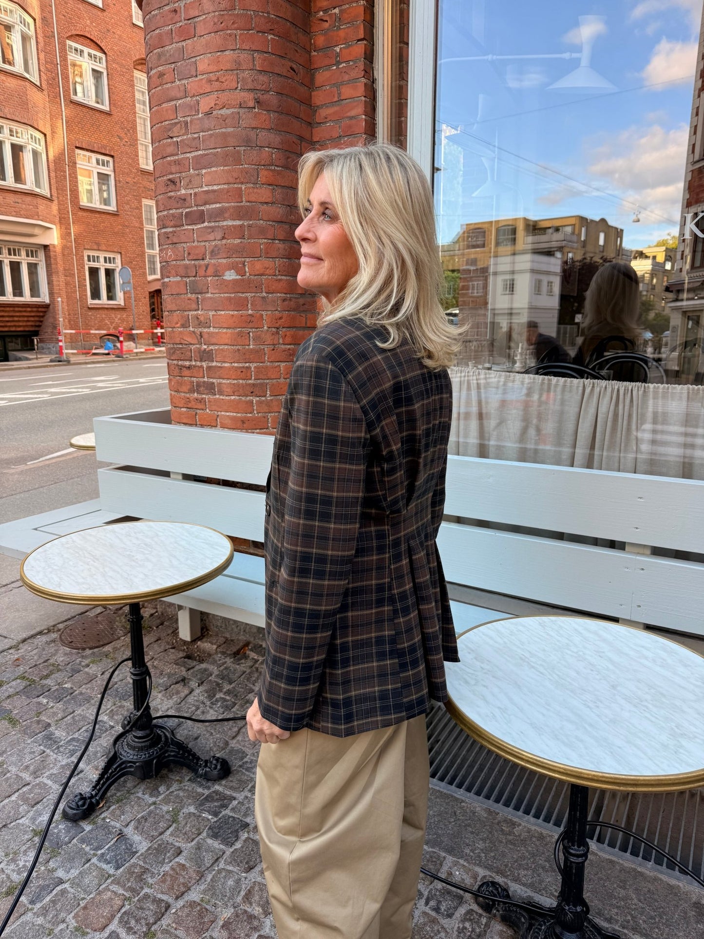 En blond kvinde står udenfor ved en hvid bænk og nogle borde iført en 2NDDAY 2NDMaeva Ternet-blazer i ternet mønster og beige bukser.