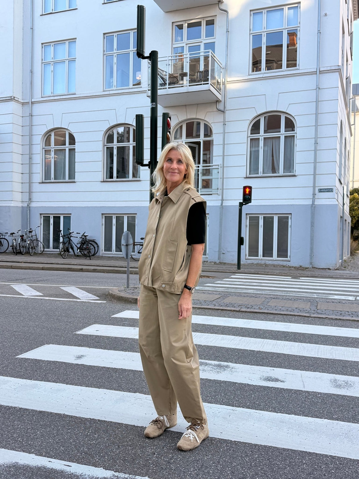 En kvinde iført bukser og vest fra 2NDDAY 2NDJimmay i farven Camel står ved et fodgængerovergang foran en hvid bygning med altaner.