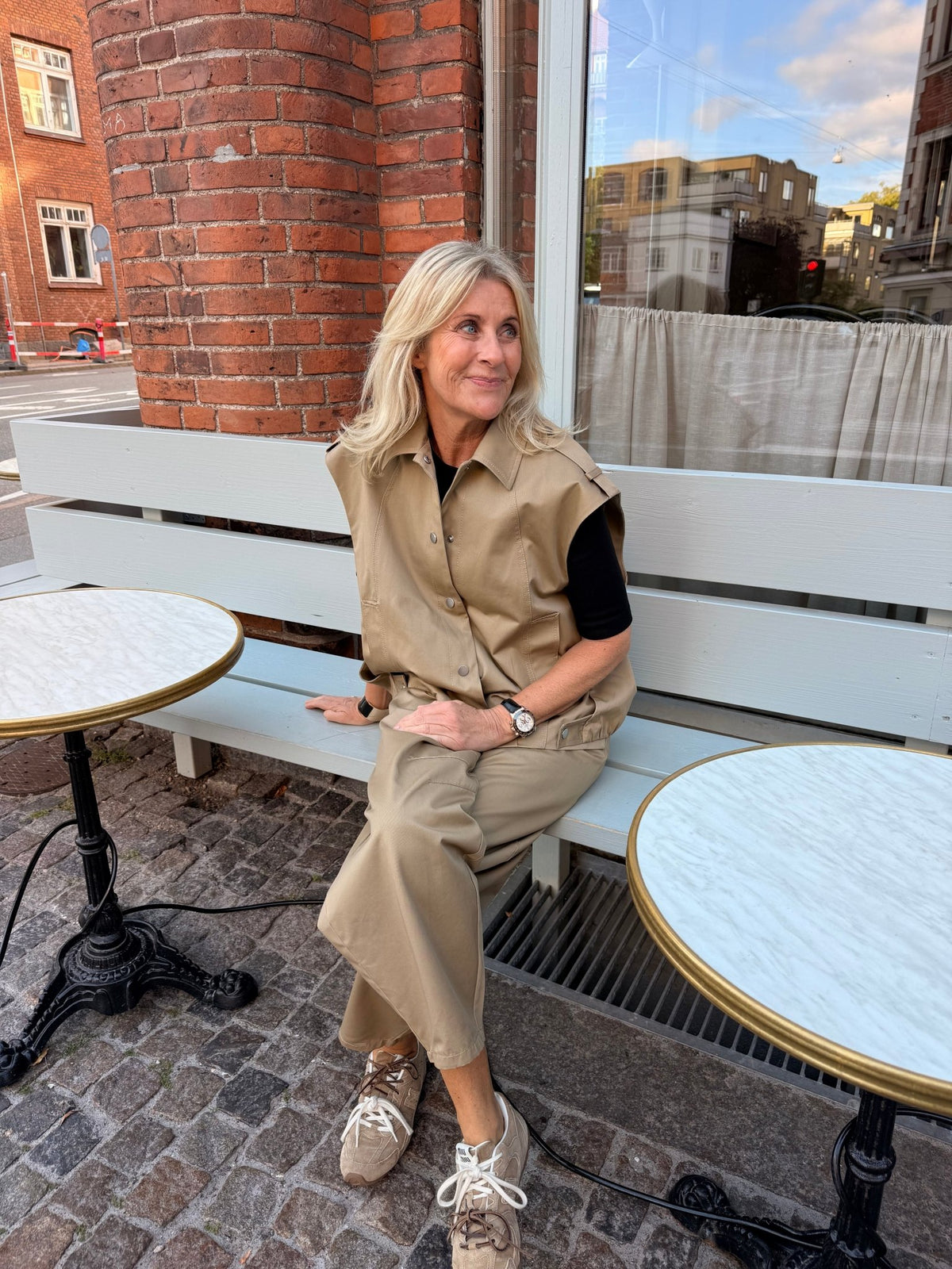 En kvinde iført en 2NDDAY 2NDCappen i farven Camel sidder på en bænk mellem to runde borde, iført sneakers, med en murstensvæg bag sig.