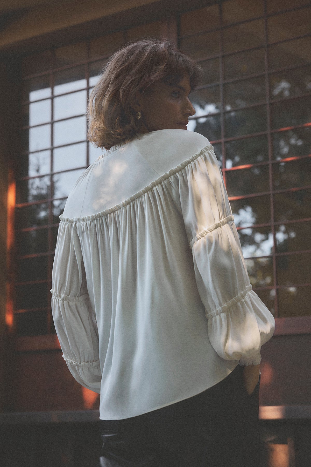 En person med kort hår har en bluse fra Gustav – Annsofie Off White 156180 på og står uden for et vindue med ruteglas.