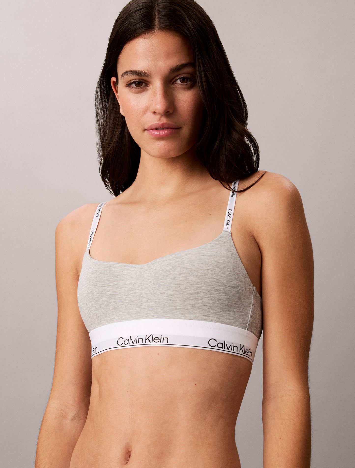 En kvinde har den lysegrå Calvin Klein QF8994-bralette med stropper med mærkelogo på og står foran en ensfarvet baggrund.