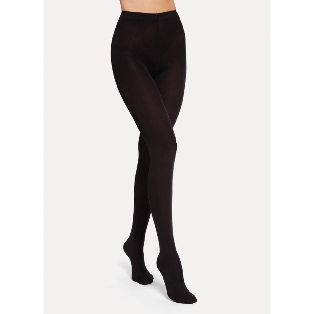 En model iført Wolford Opaque 70 Black W-strømpebukser står med det ene ben fremstrakt foran en ensfarvet lys baggrund.