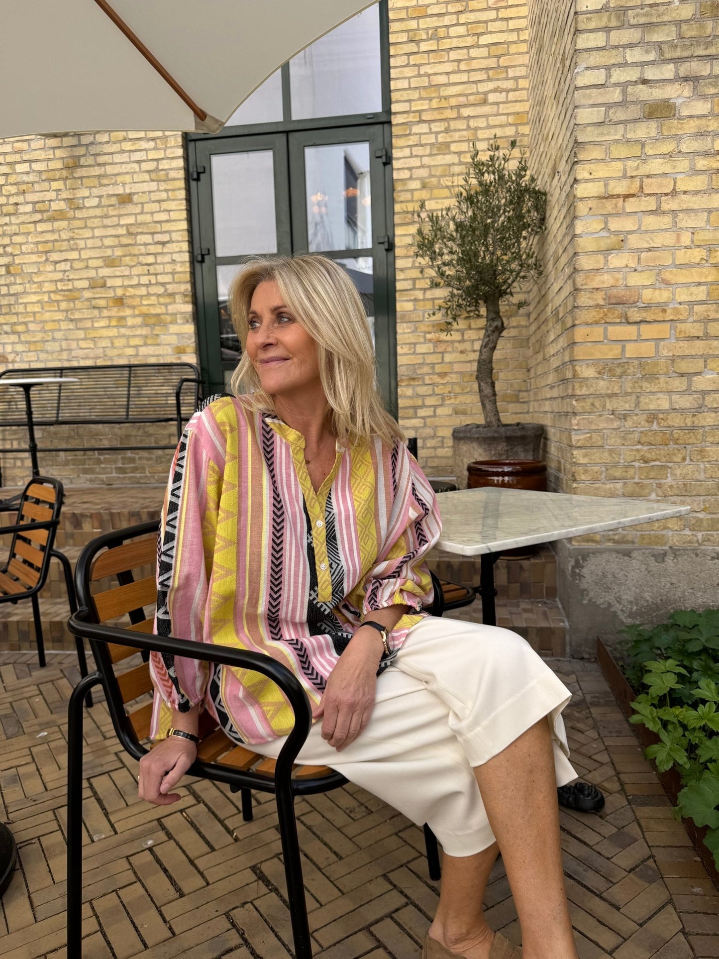 En blond kvinde i en sort Hellas-mønstret top sidder udenfor ved et terrassebord op ad en murstensvæg.