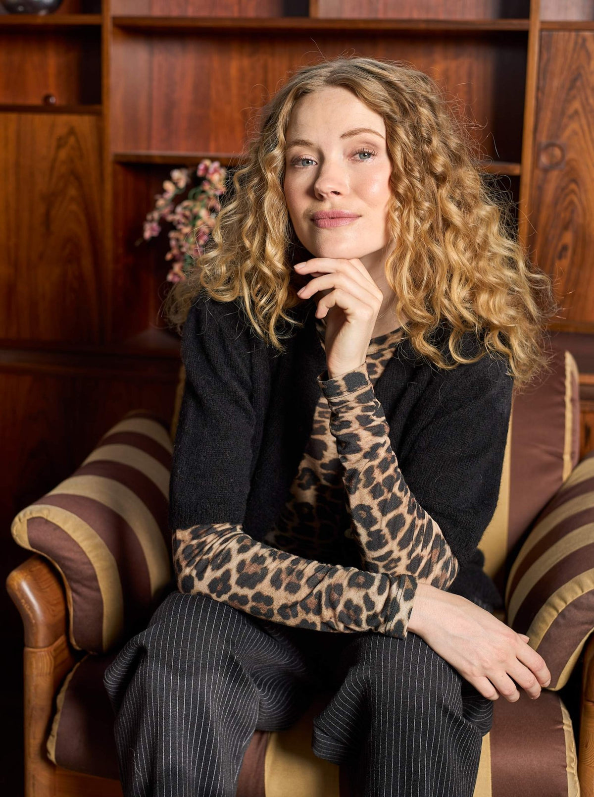 En kvinde med krøllet blondt hår sidder i en lænestol iført en Alba Leopard-bluse fra Black Colour og en sort cardigan.