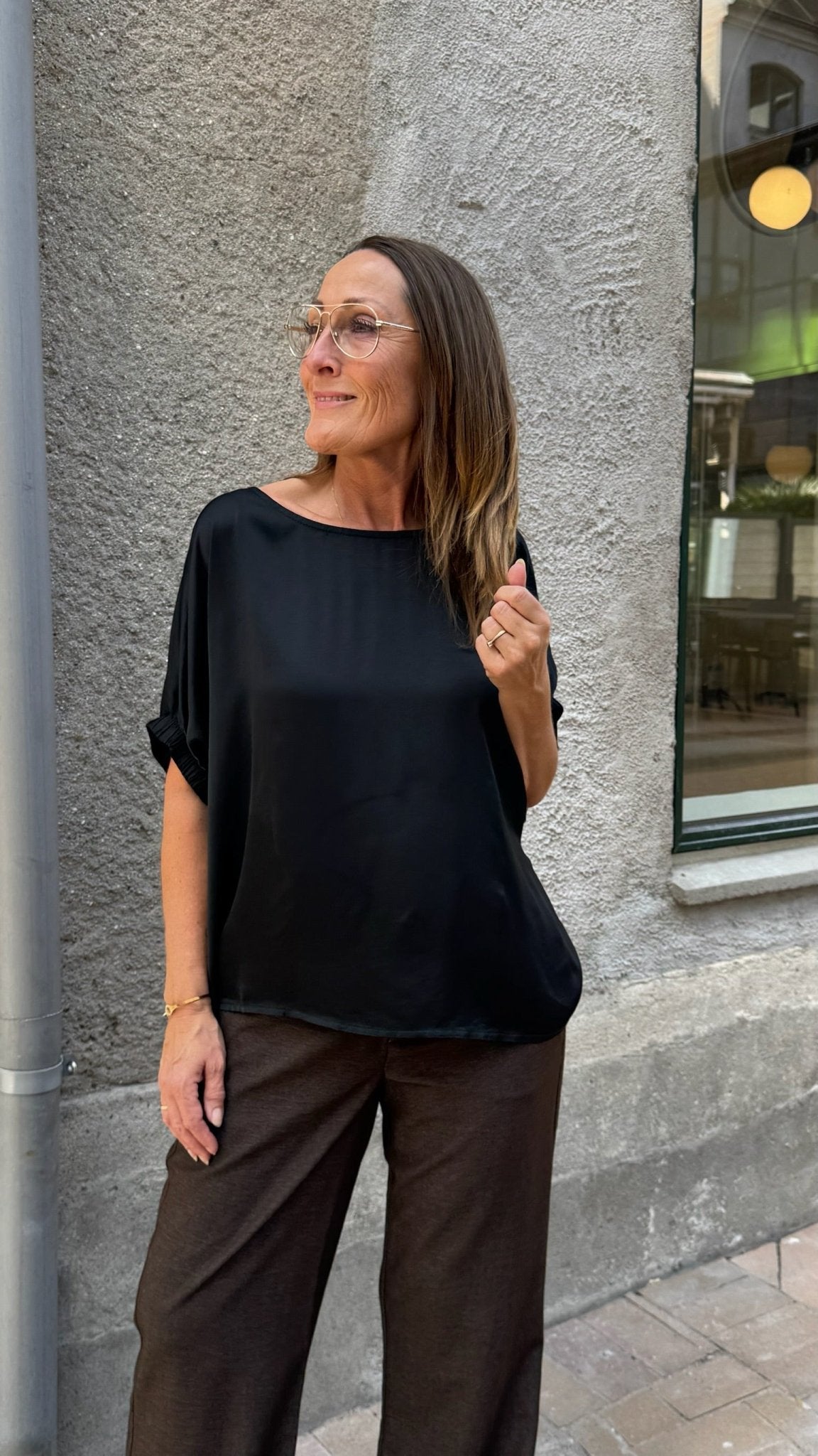 En kvinde iført en 155580 Mabel Sort-bluse fra Woman in Black Colour og brune bukser står udenfor ved en grå mur.