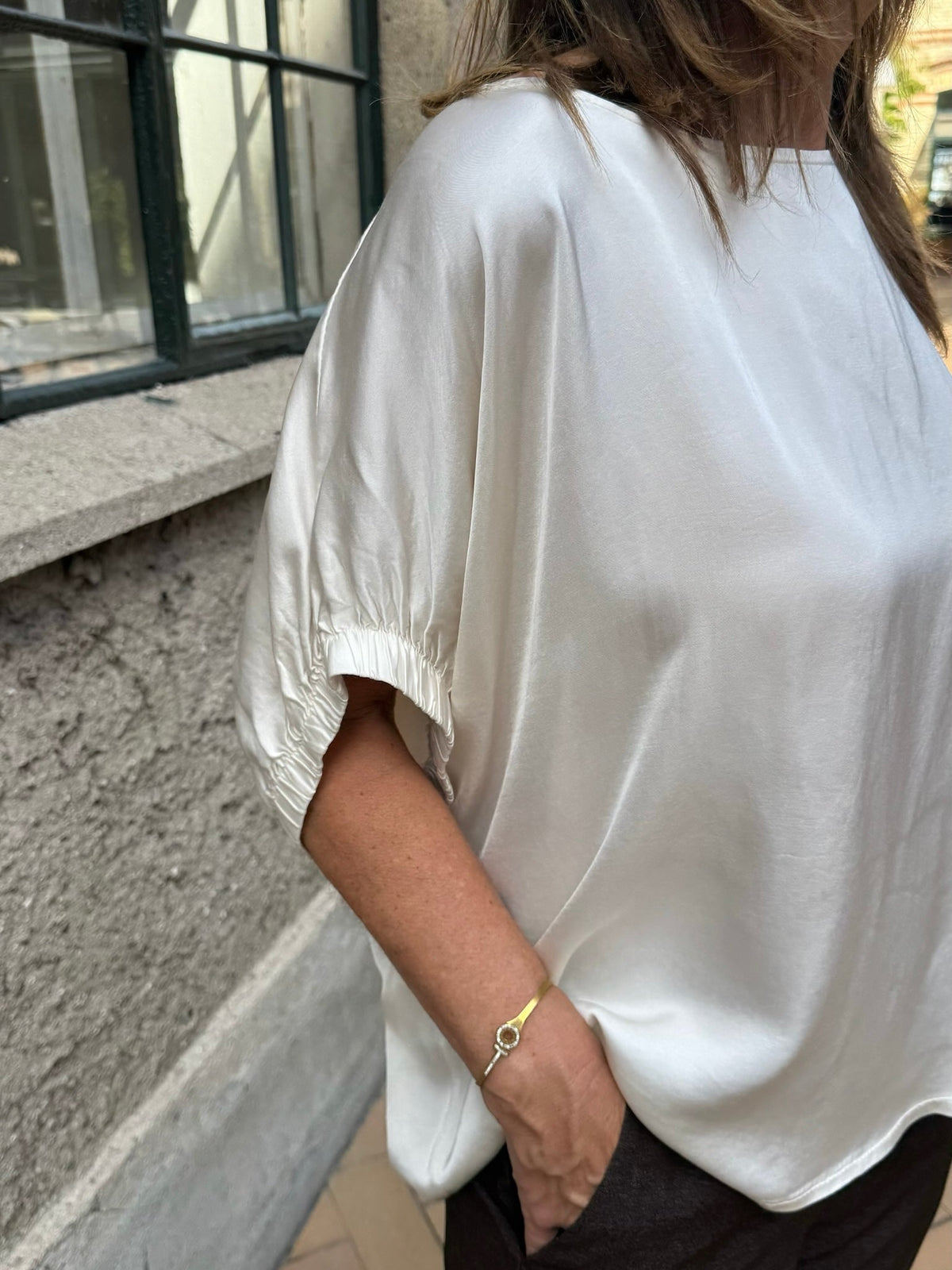 En kvinde står ved den strukturerede væg og vinduet iført en sort Mabel-bluse i off-white, mørke bukser og et guldarmbånd.