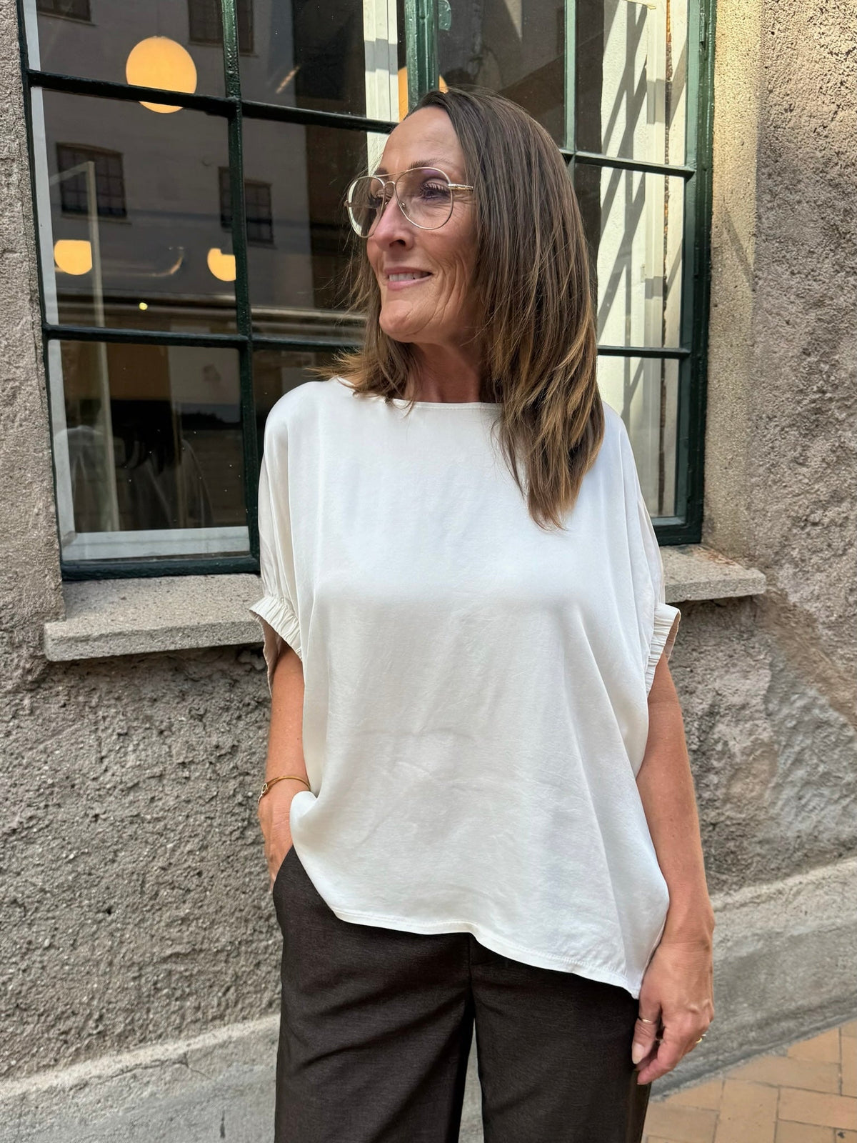 En kvinde i sort, iført en Mabel-bluse i off-white og mørke bukser, står udenfor ved en struktureret væg med et vindue.