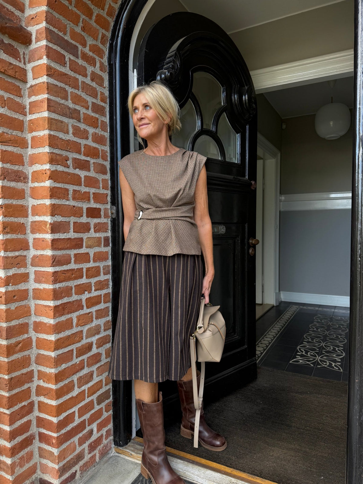 En kvinde står i en åben døråbning iført en Peta Kaffe-top fra Black Colour, stribede shorts, støvler og en beige håndtaske.