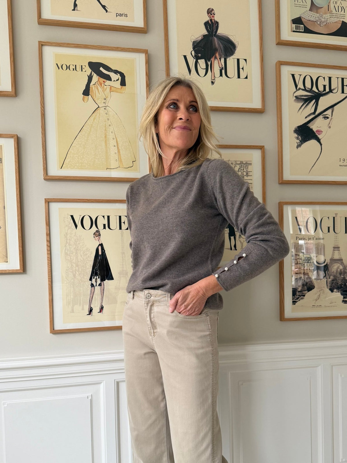 En kvinde iført en BTF CPH-kashmirbluse i brunmeleret og beige bukser står foran en væg med indrammede forsider fra magasinet Vogue.