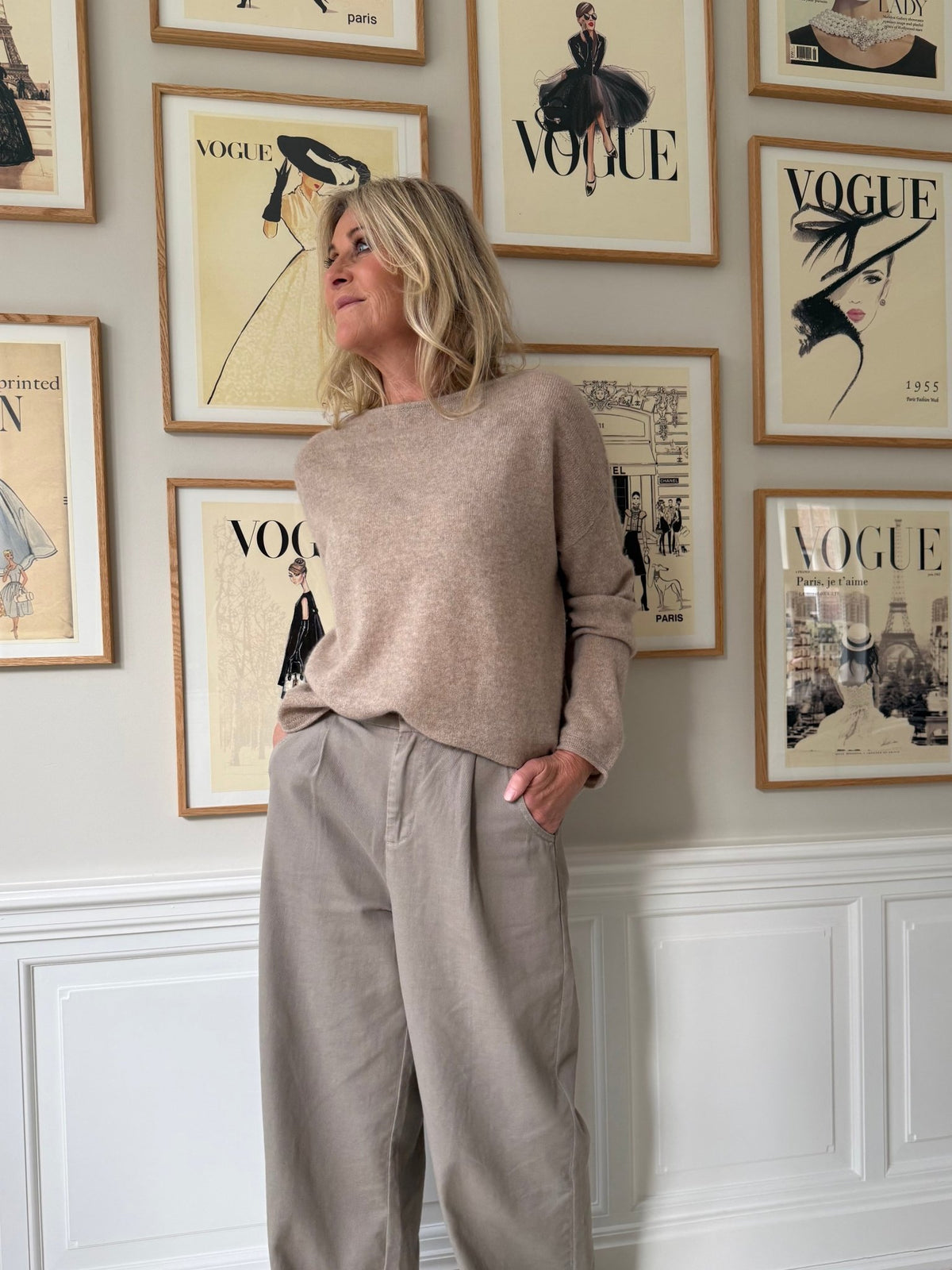 En kvinde iført en BTF CPH Cashmere Sand-trøje og taupefarvede bukser står ved en væg med indrammede forsider fra magasinet Vogue.
