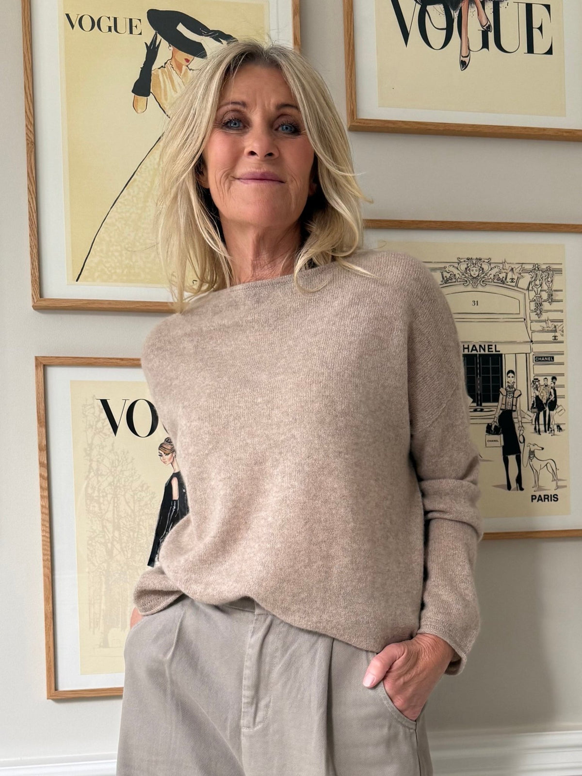 En blond kvinde iført en BTF CPH Cashmere Sand-trøje står foran indrammede forsidebilleder fra magasinet Vogue.