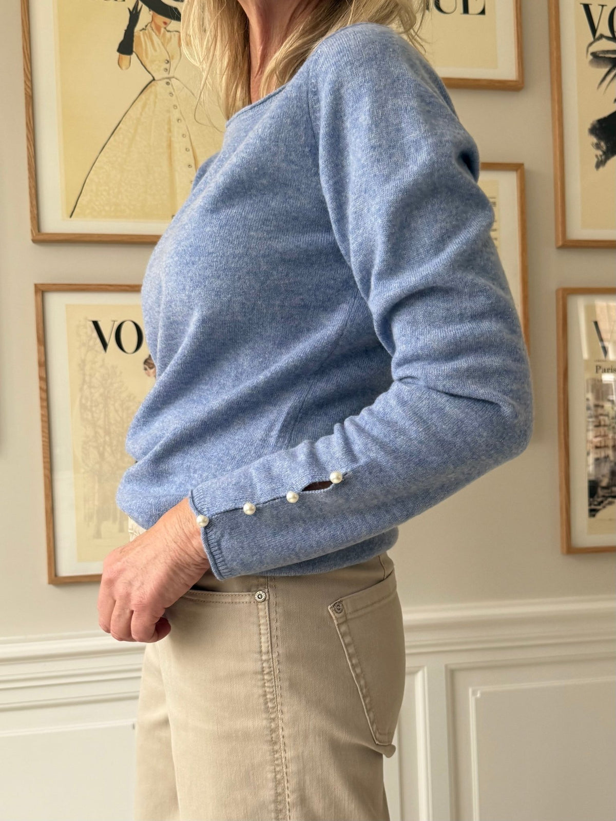 En kvinde iført en BTF CPH Cashmere Blå strikbluse med perledekorerede ærmer og beige bukser står ved siden af et indrammet kunstværk.