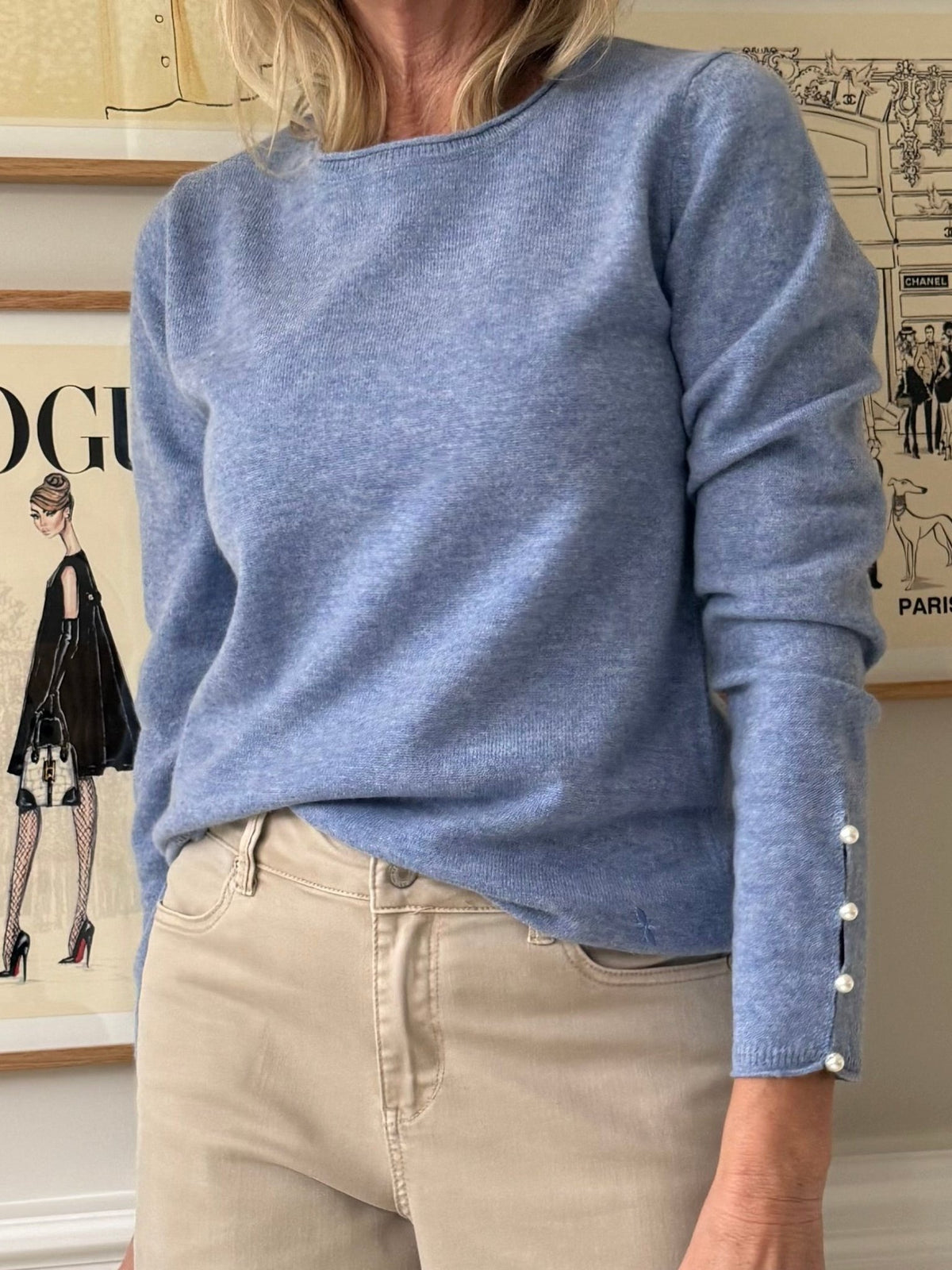 En kvinde iført en BTF CPH Cashmere Blå-strik med perlemorsfarvede ærmer og beige bukser står indendørs foran et indrammet kunstværk.