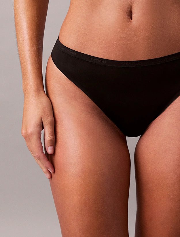 En person iført sorte Calvin Klein Basic 2025-trusser i brasiliansk snit, der står med den ene arm afslappet mod en ensfarvet baggrund.