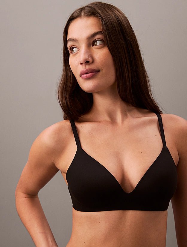 En kvinde med langt brunt hår viser Calvin Klein 150566 Wire Free Sort-bh’en frem foran en grå baggrund, mens hun kigger til siden.