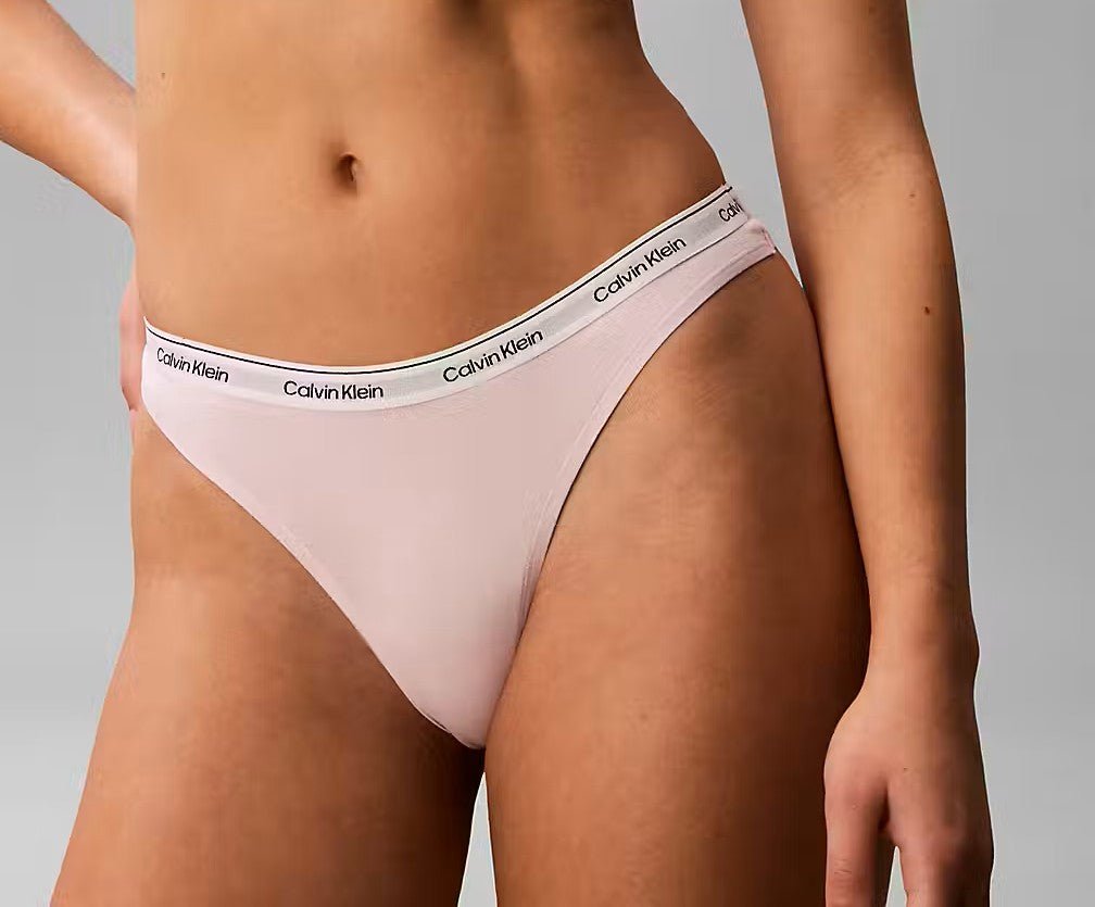 Model iført lyserød Calvin Klein 153667 G-streng, logo i linningen, overkrop og ben afskåret, grå baggrund.