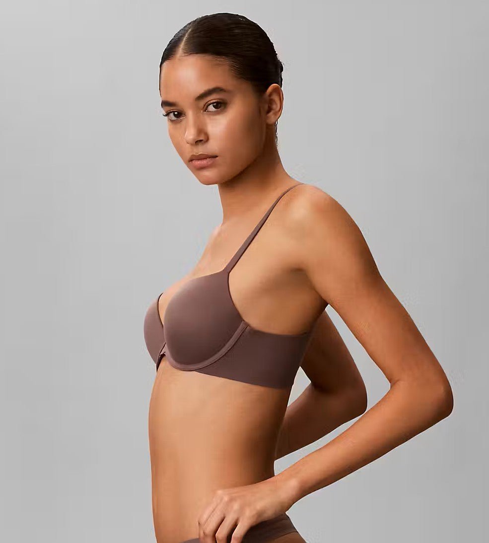 Model iført Calvin Klein 155200 Botanical Brown Brun-bh og -trusser, der poserer i profil foran en ensfarvet grå baggrund.