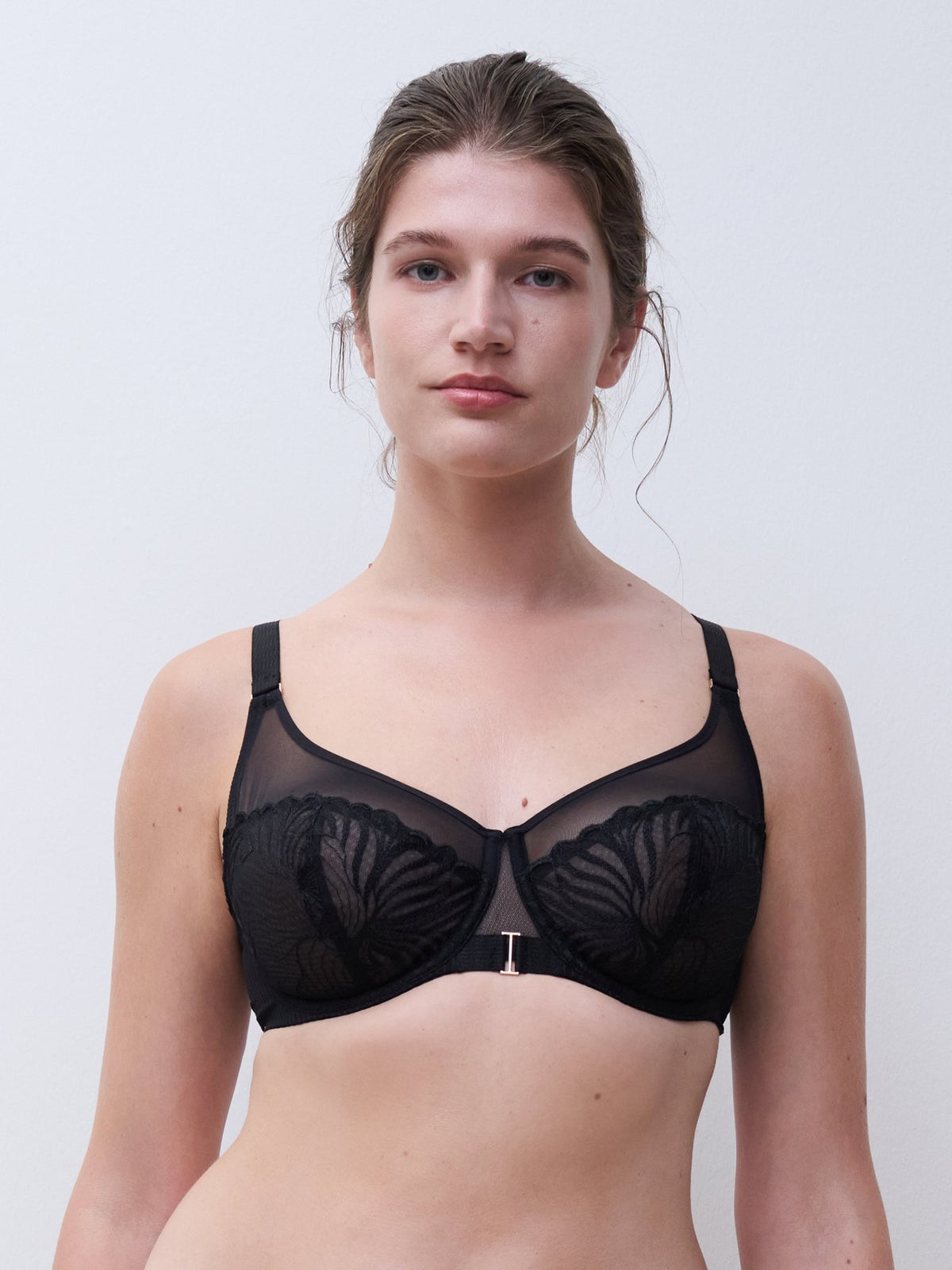 Kvinde iført en Chantelle – Legend Black Sort-bh med blomstret netstof, stående foran en ensfarvet lys baggrund.