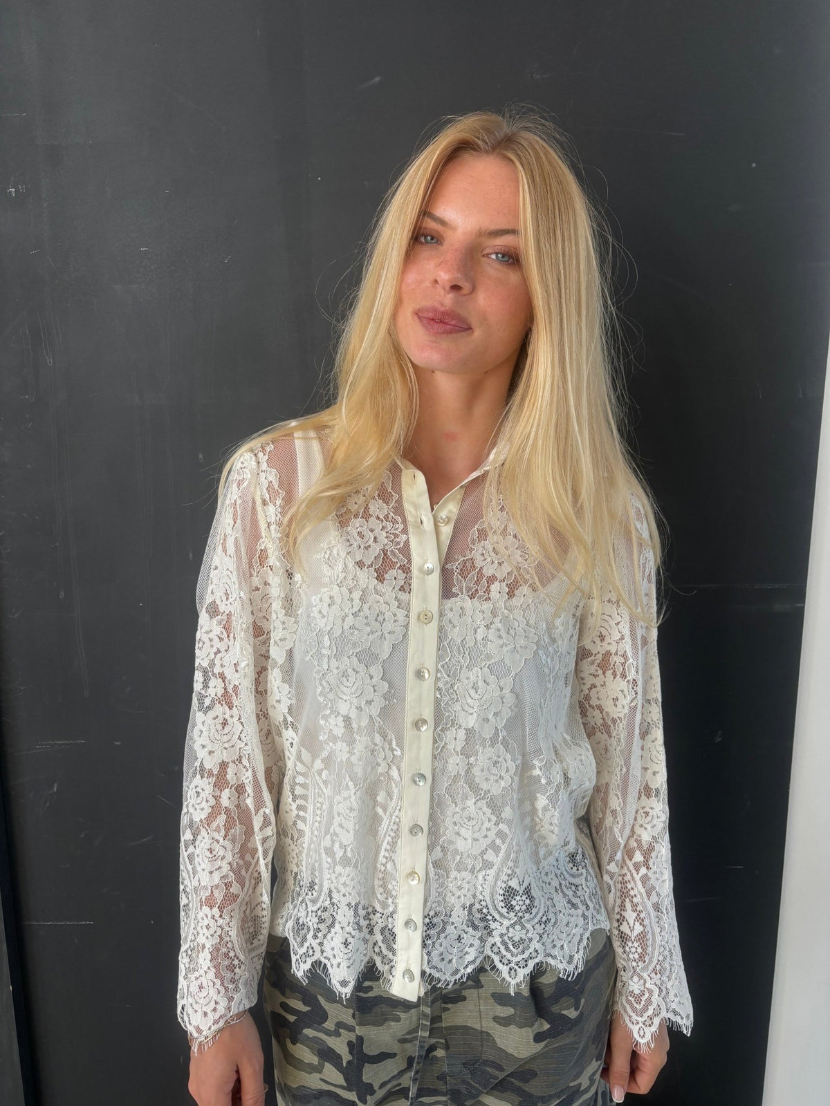 En blond kvinde poserer foran en sort baggrund iført en CONTINUE - Paloma Off hvid skjorte fra CONTINUE og camouflagebukser.
