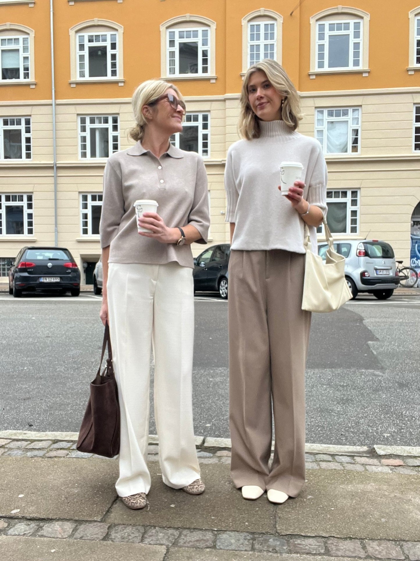 To kvinder i afdæmpede outfits, der begge har Copenhagen Muse Tailor-bukser i farven »Off-white« på, står på et fortov i byen med en kop kaffe.