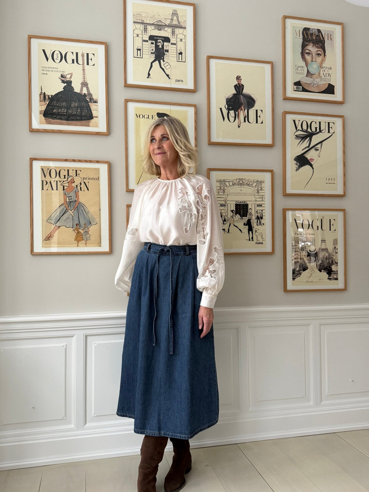 En kvinde i en Muse Molly Off White-bluse i hvid og en denimnederdel står ved en væg med gamle Vogue-forsider.