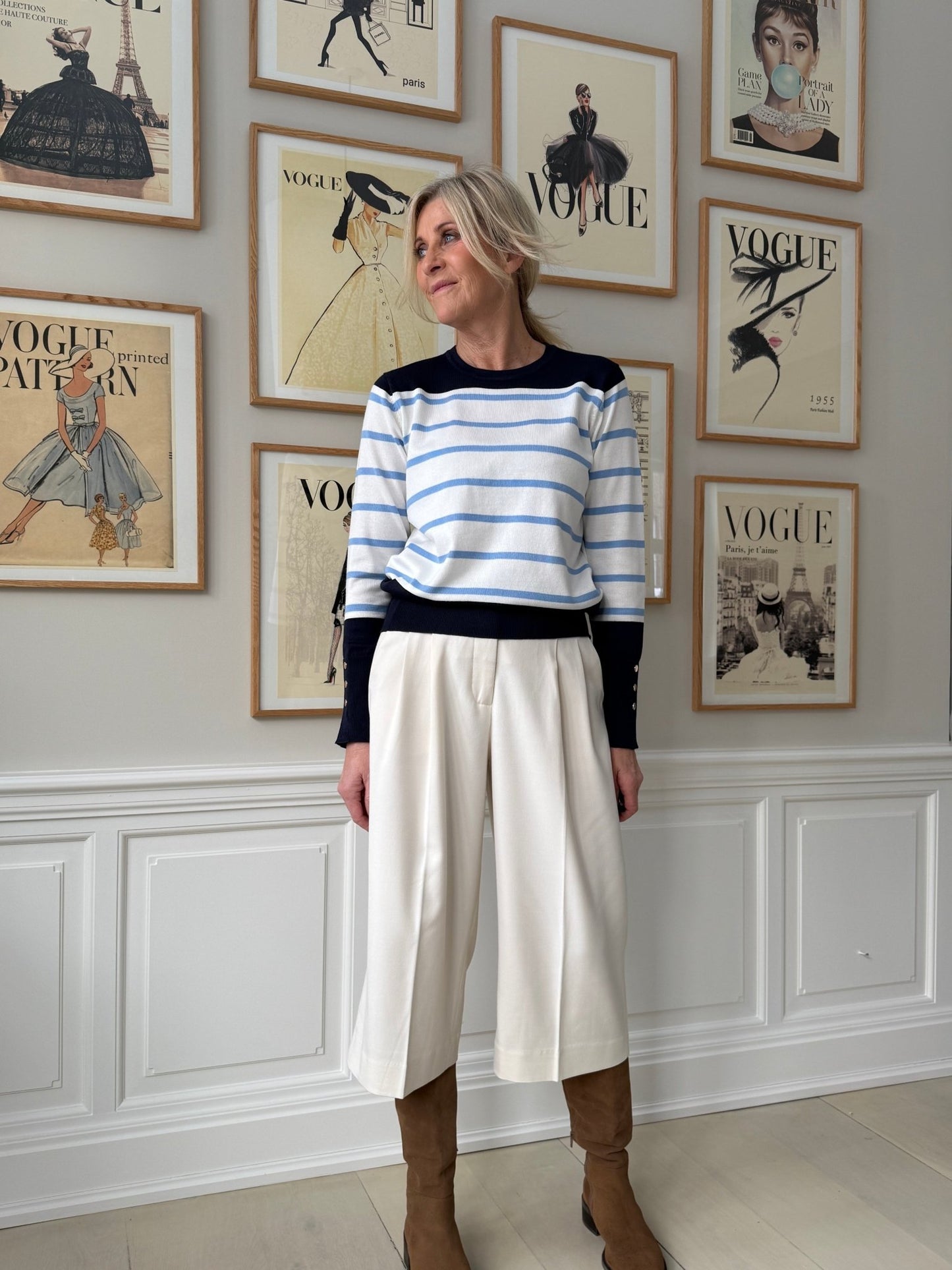 En kvinde i culottes i farven »Off White« fra Muse Tailor i København står foran indrammede Vogue-forsider, iført en stribet sweater.