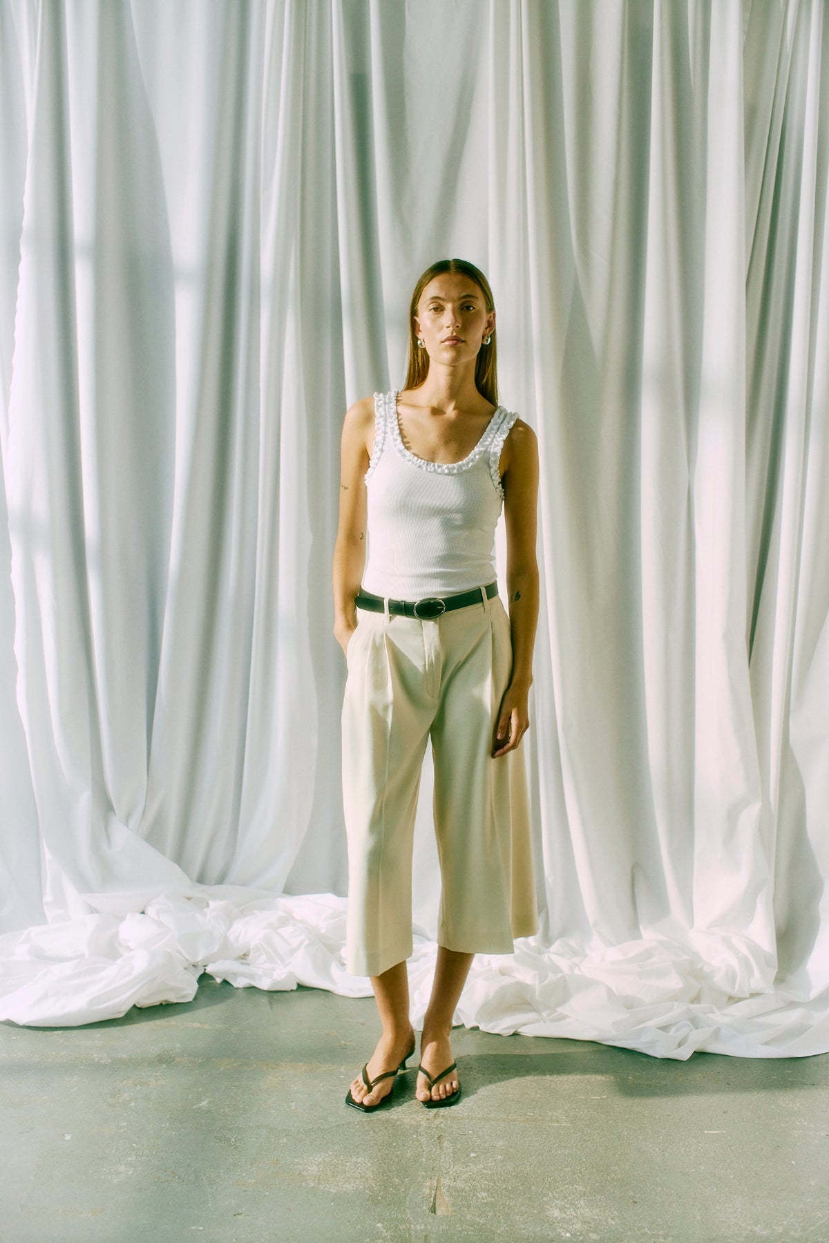 Kvinden foran de hvide gardiner har en offwhite tanktop fra Copenhagen Muse Tailor, et sort bælte og sandaler på.