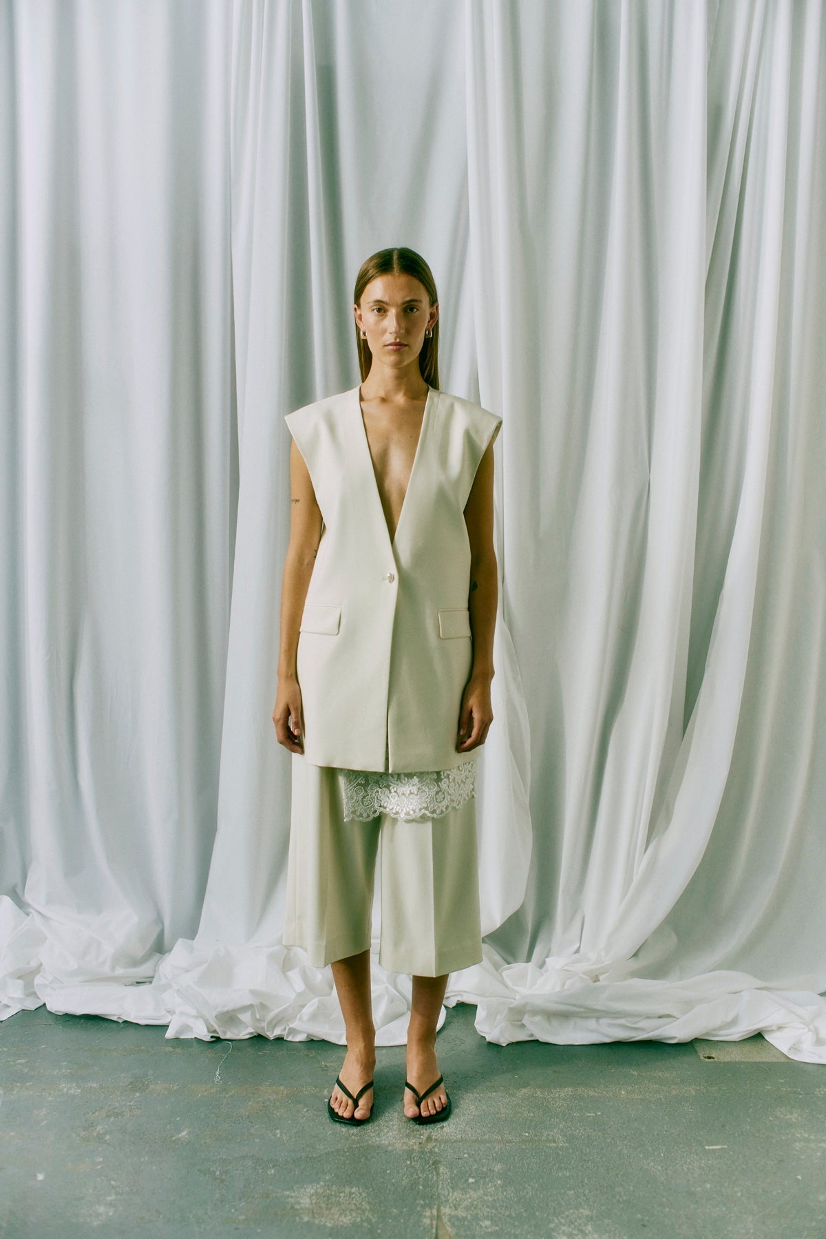 En kvinde foran hvide gardiner iført bermudashorts fra Copenhagen Muse Tailor i farven »Off White« og en cremefarvet vest samt sorte sandaler.