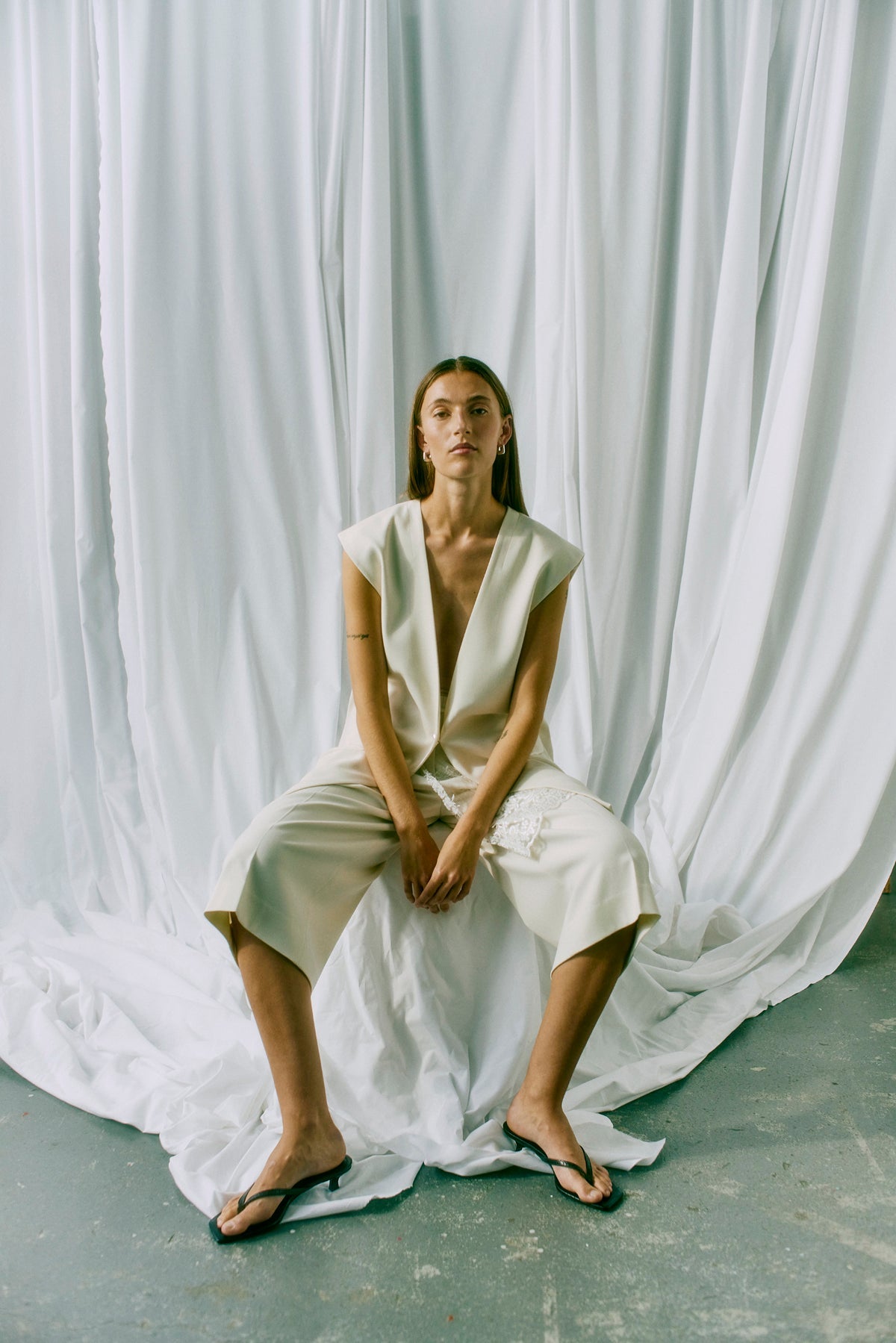 En kvinde i København iført offwhite bukser og sandaler fra Muse Tailor sidder på en stol med hvidt, draperet stof i baggrunden.