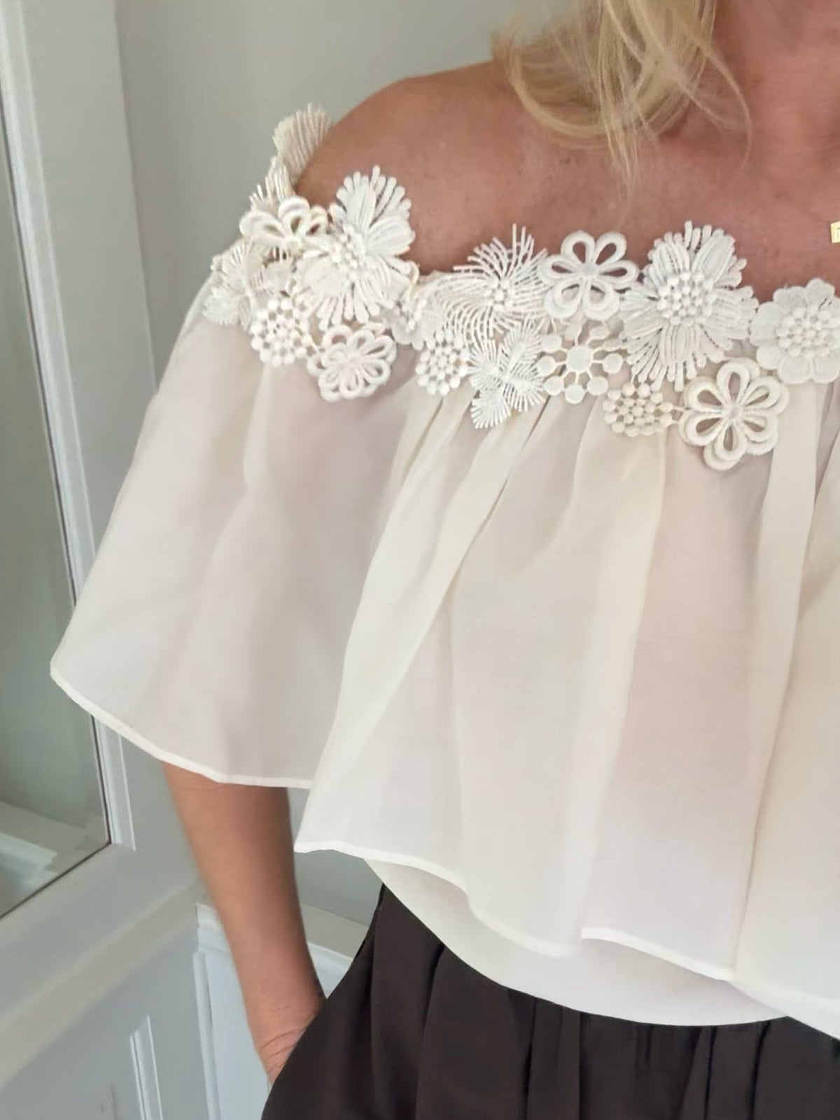 Kvinde i København. Muse Molly Off-White-bluse med bare skuldre og blondekant samt sorte bukser, stående indendørs.