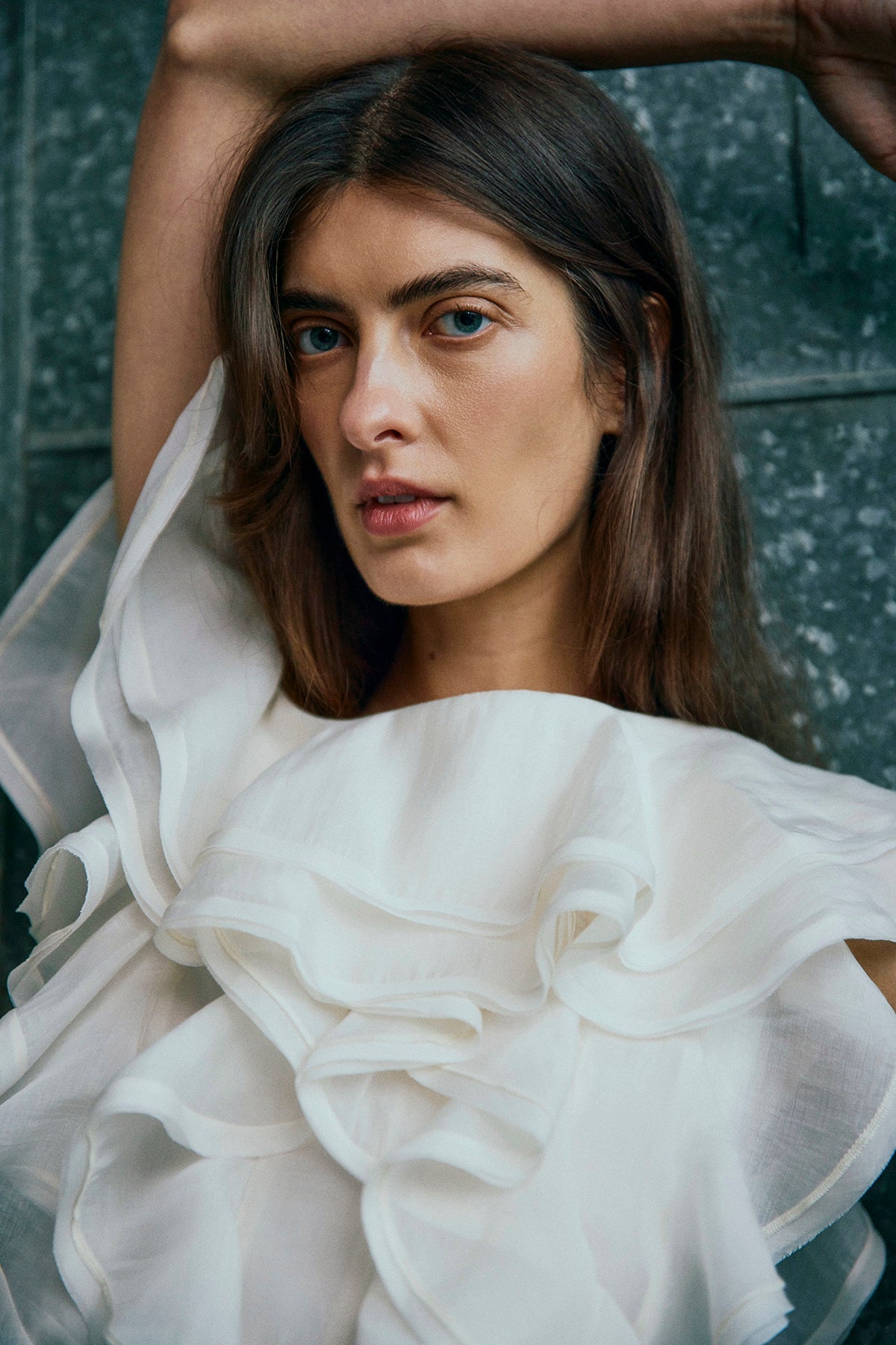 En kvinde med langt brunt hår har en offwhite Tilly-bluse fra Copenhagen Muse på og poserer foran en struktureret grå baggrund.