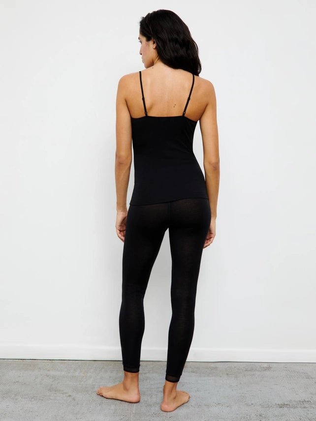 En kvinde iført FEMILET Juliana Sort-leggings og en camisole står barfodet på et tæppe med ansigtet vendt mod en ensfarvet hvid væg.